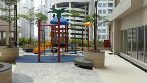An outdoor playground with slides and climbing structures, surrounded by potted plants and a building in the background.