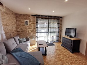 A living room with a stone wall, a grey sofa, a wooden coffee table, a TV on a wooden stand, and a large window with curtains. - Gîte Corrézien 3 pers. Heated swimming pool (Chamboulive)