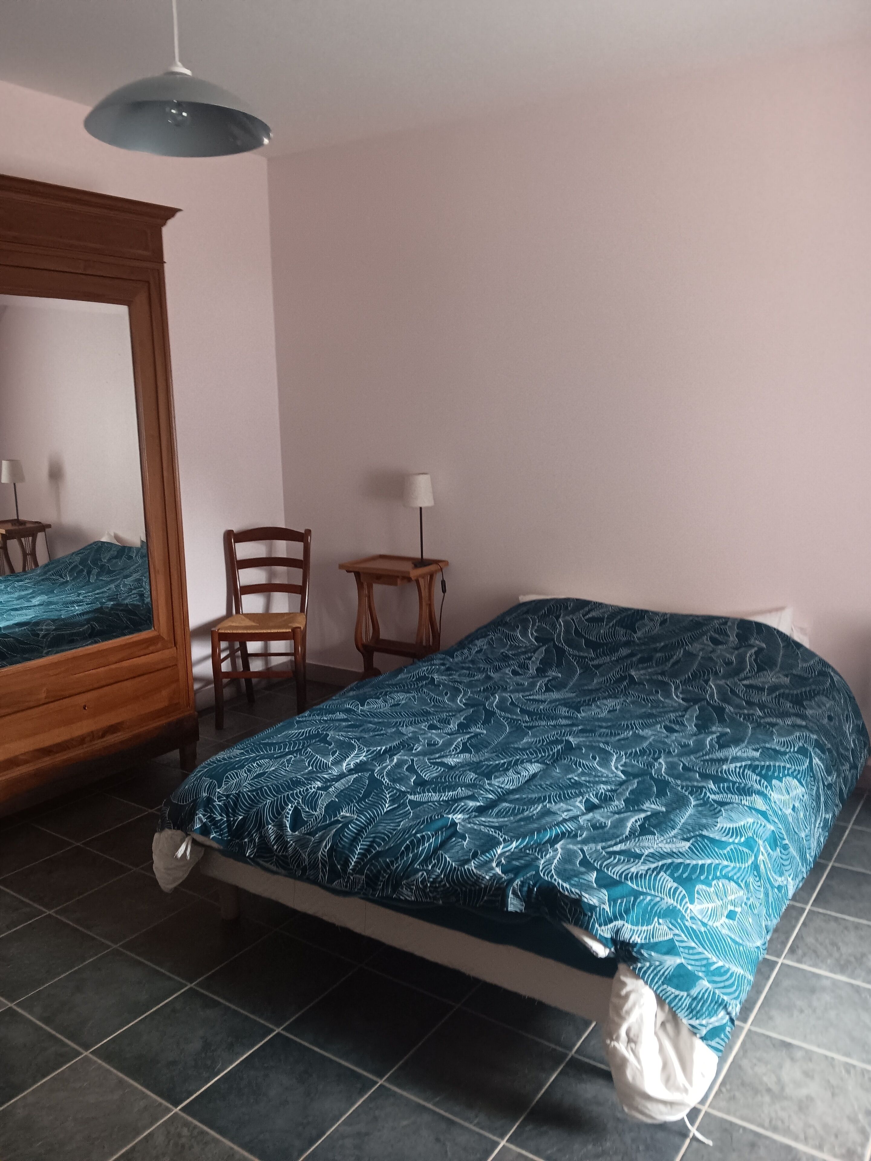 Un dormitorio con una cama, una silla, una mesita, una lámpara y un espejo.