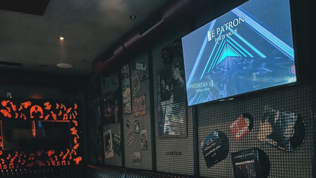 A restaurant interior with a long wooden table, a booth seating area, and a screen displaying 'The Patron Party Mode'. - Le Patron Hotel (Lahr)
