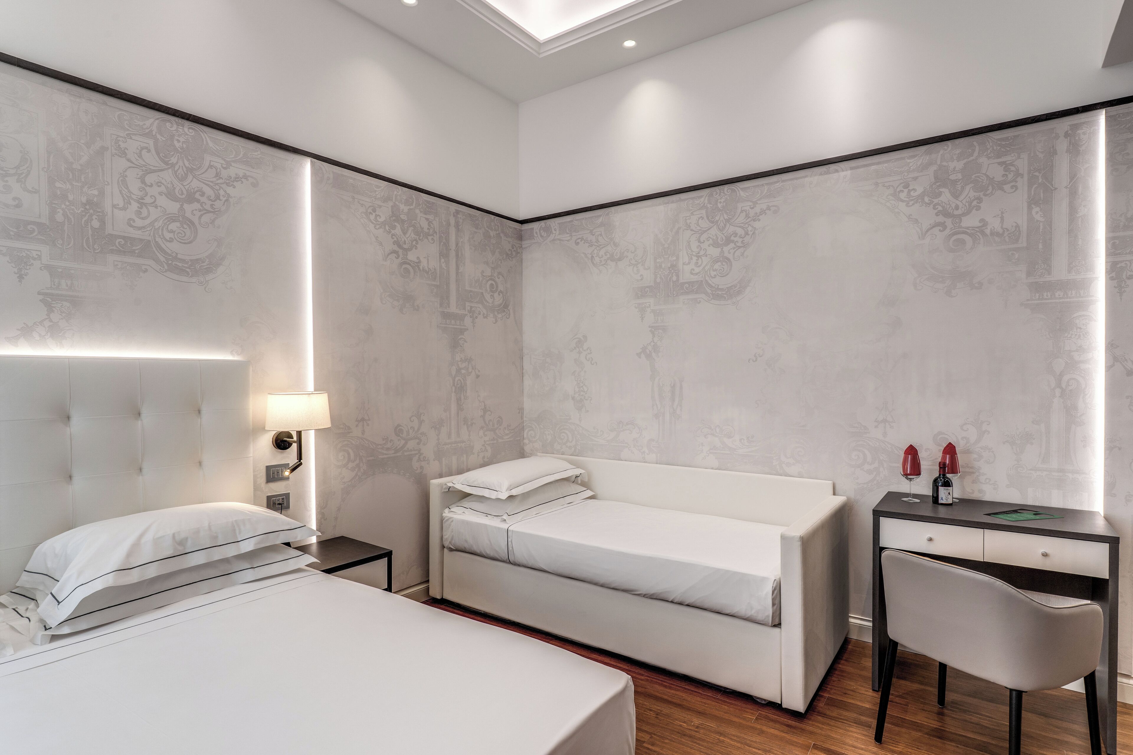 A modern hotel room with a large bed, a smaller bed, a desk with a chair, and a nightstand with a lamp.