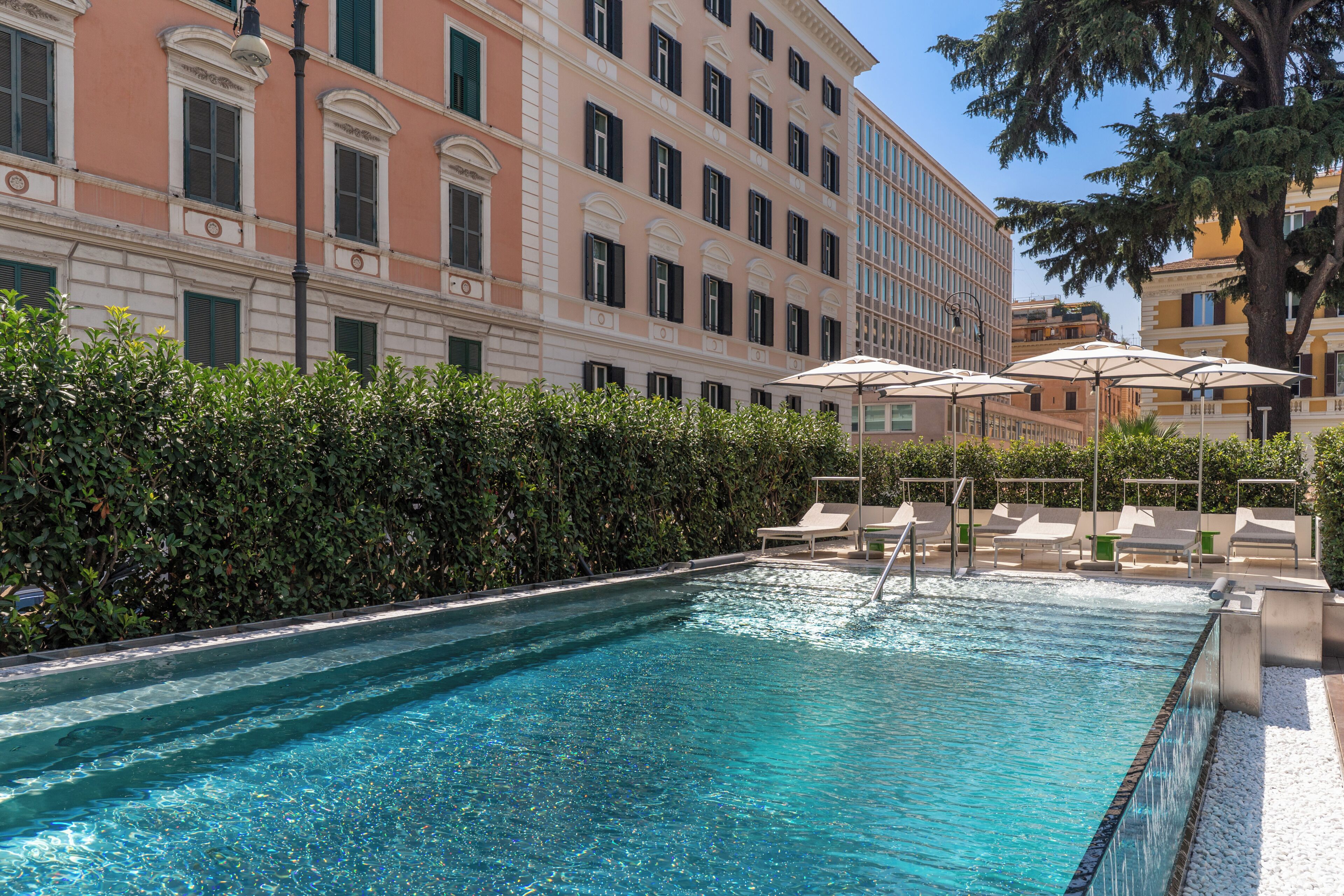 Una piscina con lettini e ombrelloni, circondata da un siepe e un edificio con diverse finestre.