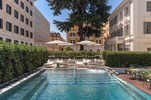 A swimming pool with lounge chairs and umbrellas, surrounded by hedges and buildings.