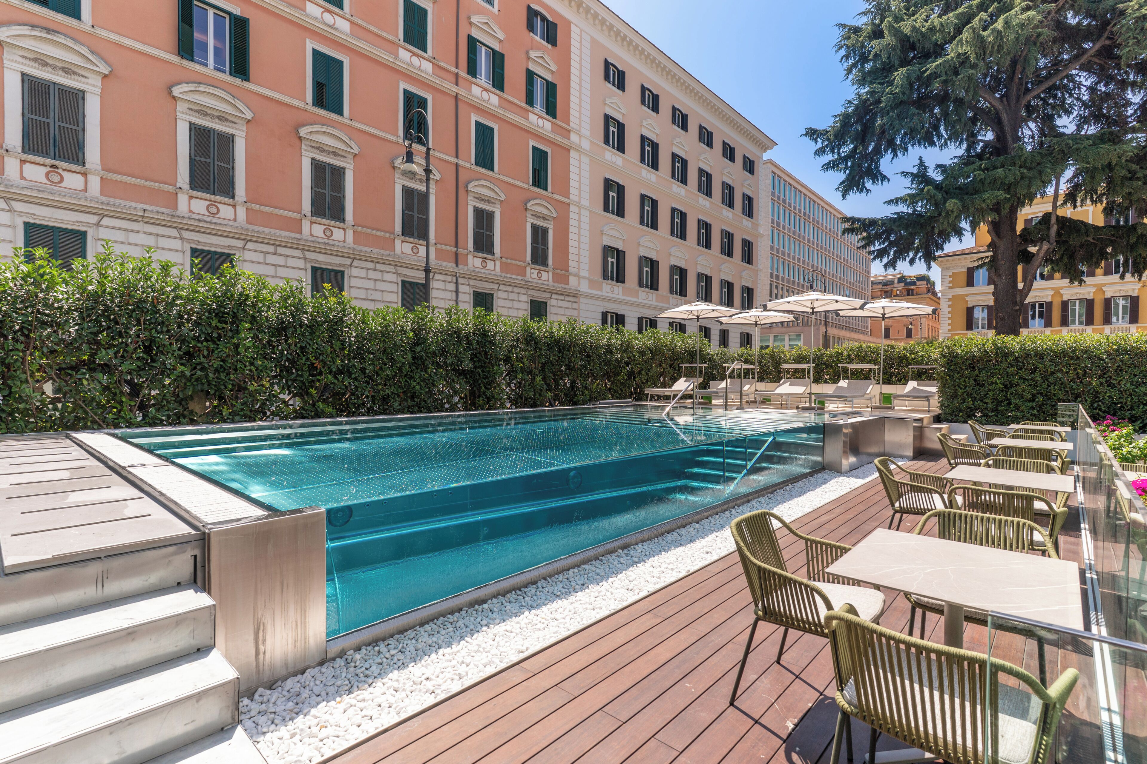 Una piscina con cubierta de madera, rodeada de vegetación y un edificio con múltiples ventanas.