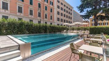 Una piscina con cubierta de madera, rodeada de vegetación y un edificio con múltiples ventanas.