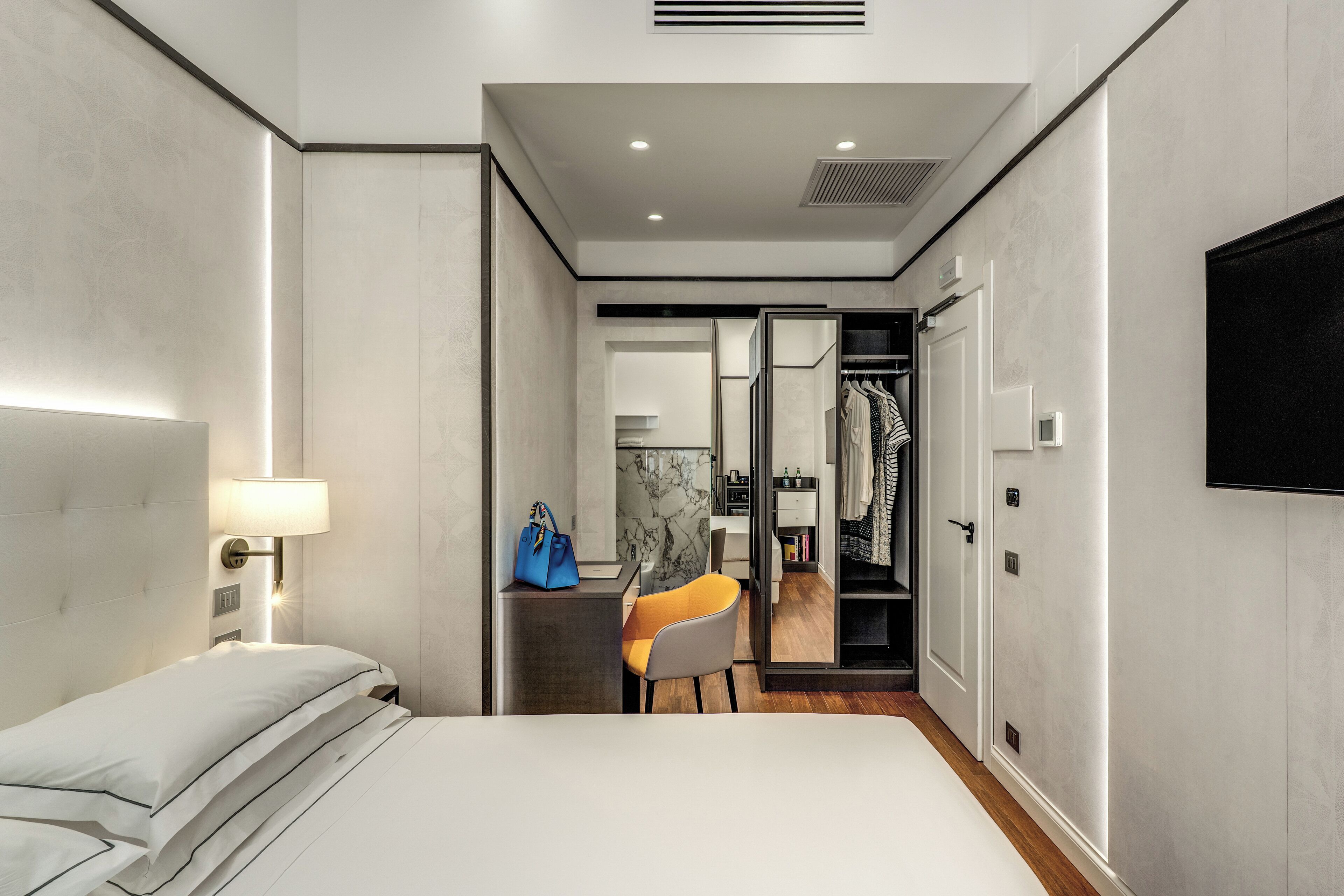 A modern hotel room with a bed, a desk, and a chair. There is a wardrobe with a mirror and a TV mounted on the wall.