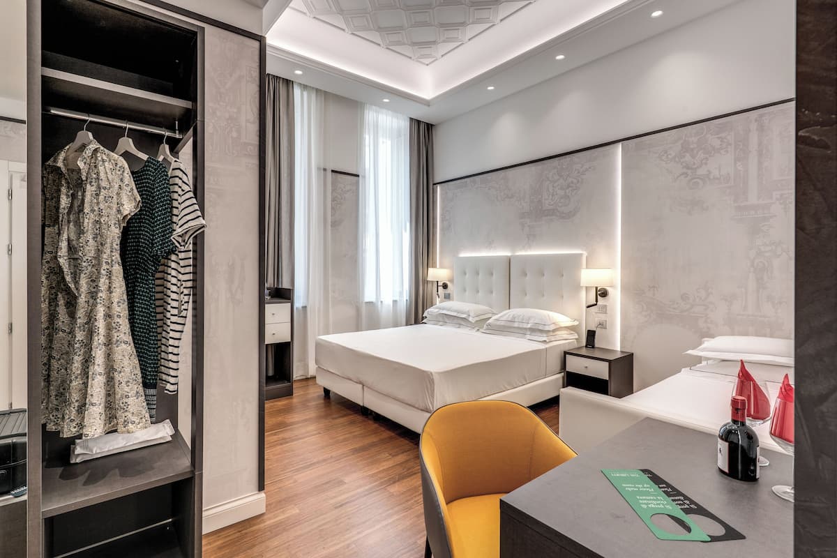 A modern hotel room with a neatly made bed, a wardrobe with hanging clothes, a desk with a bottle and glasses, and a bright yellow chair.