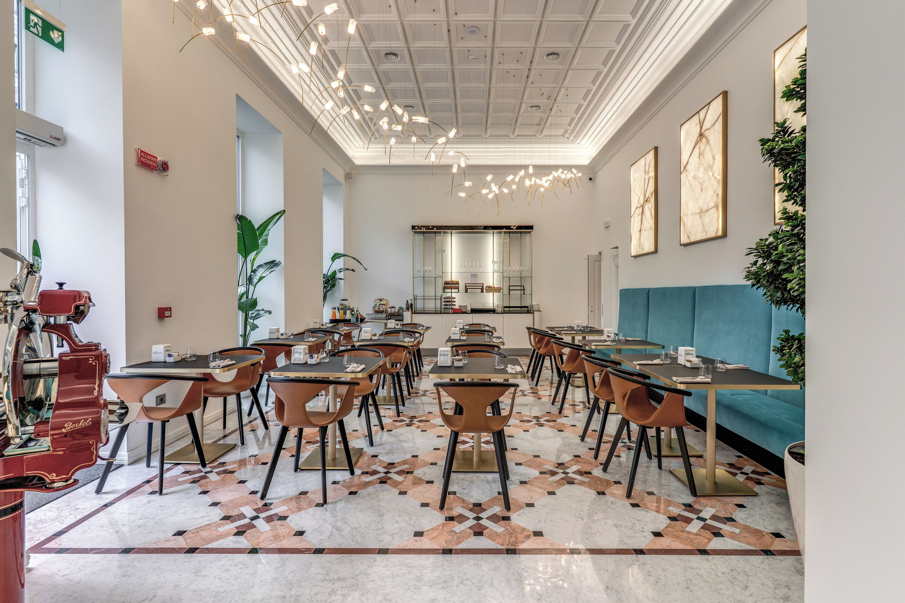 A hotel dining area with multiple tables and chairs, a kitchen in the background, and a patterned floor.