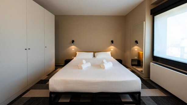Una habitación de hotel moderna con una cama grande, dos mesitas de noche, un armario y un amplio ventanal.