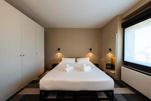 A modern hotel room with a large bed, two bedside tables, a wardrobe, and a large window.