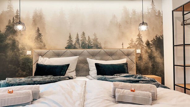 Une chambre à coucher avec une tête de lit capitonnée, une fresque murale sur le thème de la forêt, deux lampes suspendues et des serviettes soigneusement rangées.