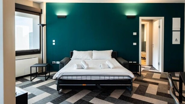 A modern hotel room with a large bed, a small table, and a chair. There is a window providing natural light and a door leading to another room.