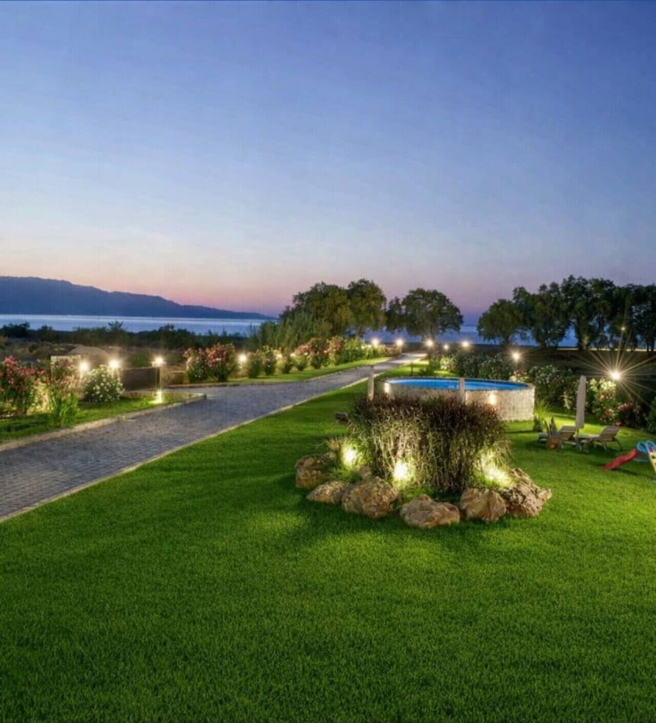 A well-lit outdoor area with a pathway leading to a pool, surrounded by lush greenery and a view of the water.