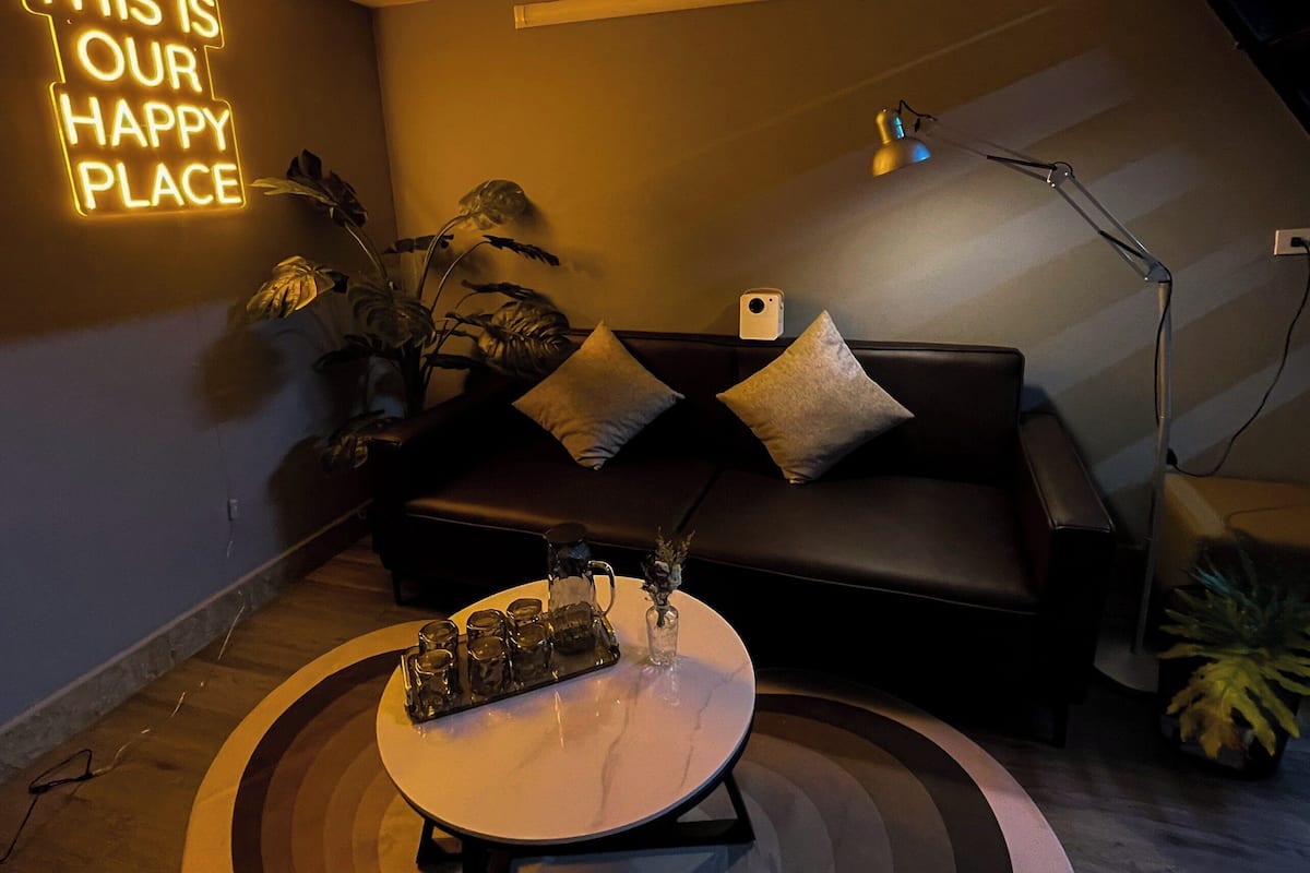 A modern living room with a black leather sofa, a coffee table with glasses, a neon sign, and a plant.