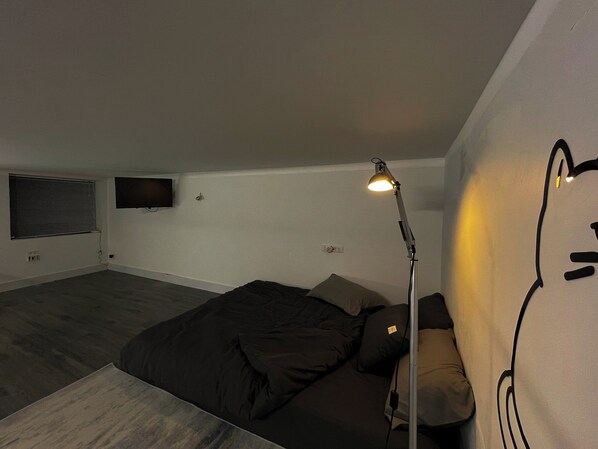 Un dormitorio con cama, lámpara de escritorio, televisor montado en la pared y ventana con persianas.