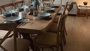 Een eethoek met een lange houten tafel, gedekt voor zes personen, omringd door houten stoelen. Op tafel staat een vaas met gedroogde planten, en de kamer heeft een moderne uitstraling met een flatscreen televisie aan de muur.