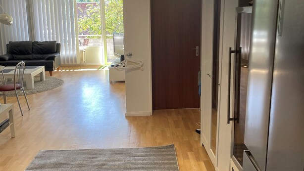 Un vaste espace de vie agrémenté d’un grand tapis gris, d’un canapé noir et d’une table à manger. Une cuisine moderne avec un réfrigérateur en acier inoxydable est visible sur la droite.