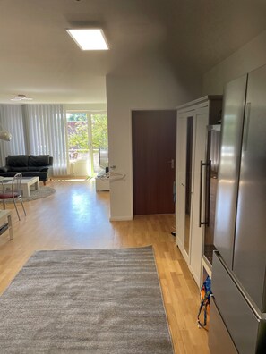 A spacious living area with a large grey rug, a black sofa, and a dining table. A modern kitchen with a stainless steel refrigerator is visible to the right. - Furnished 3-room apartment with balcony in the hotspot of Hamburg-Eimsbüttel. (Hamburg)