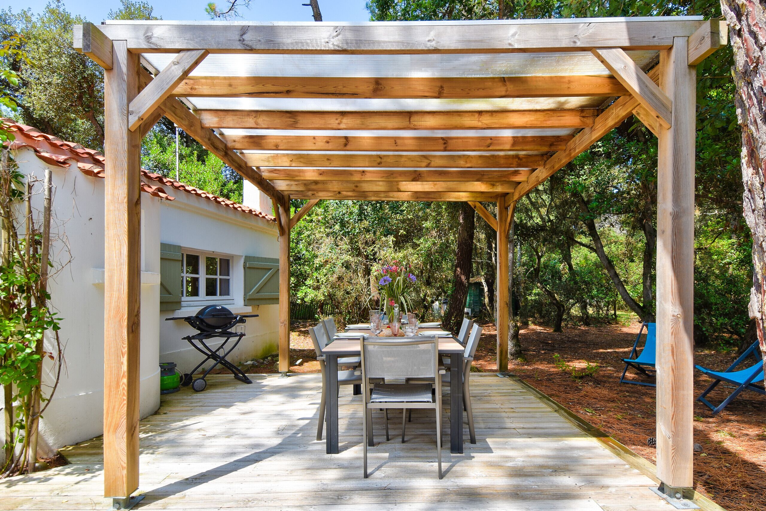 Eine Holzupergola mit Tisch und Stühlen darunter, umgeben von Bäumen und einem kleinen Gebäude mit Ziegeldach.