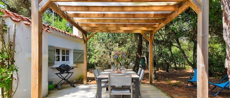 Eine Holzupergola mit Tisch und Stühlen darunter, umgeben von Bäumen und einem kleinen Gebäude mit Ziegeldach.