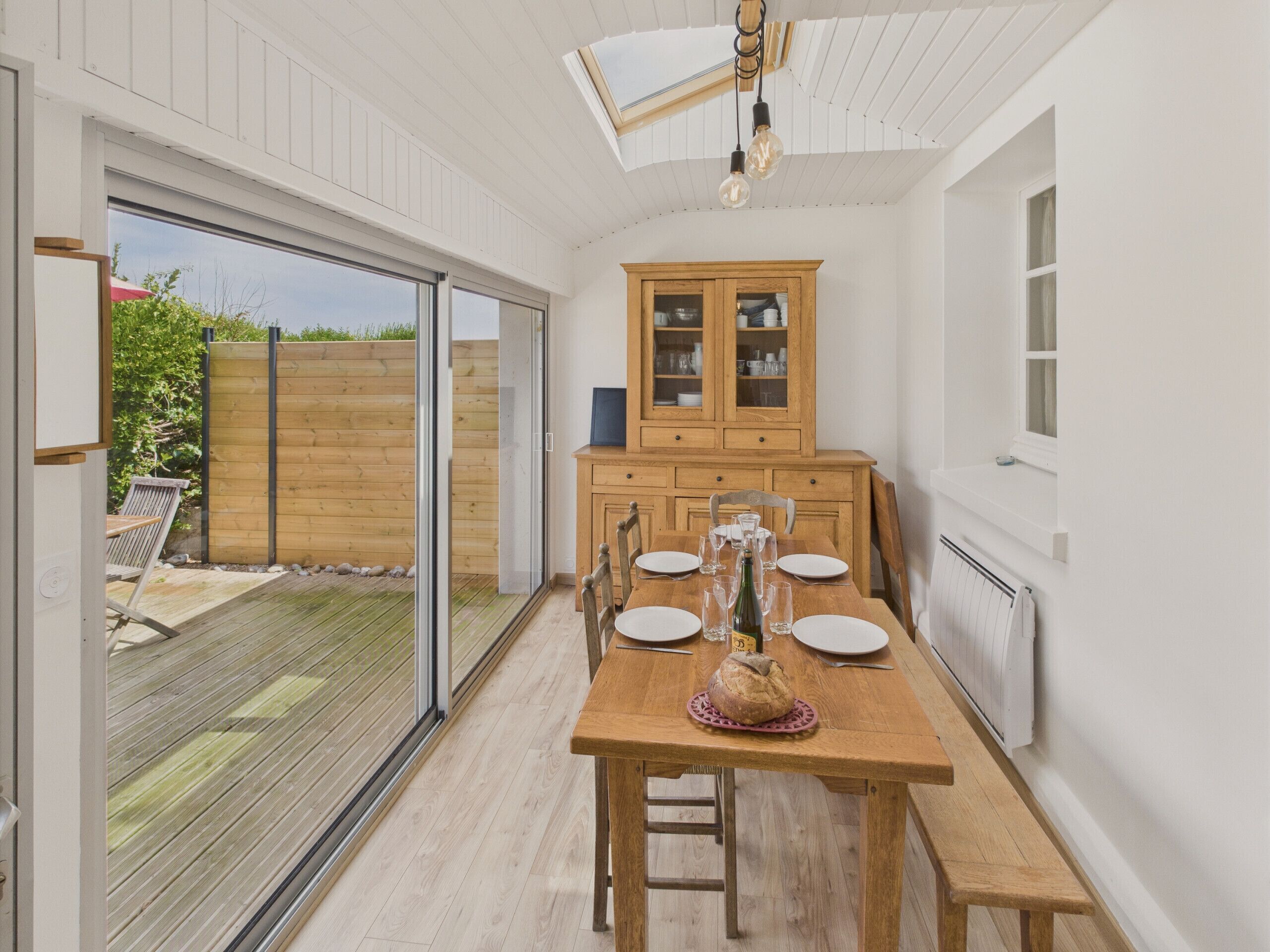 A kitchen with a wooden table set for dining, a cabinet with glass doors, and a view of the outdoor garden through a sliding glass door.