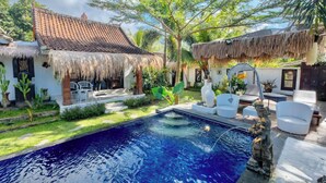 Uma área de piscina com uma casa de telhado de palha, móveis de exterior brancos e plantas tropicais.