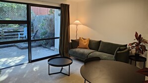 Ein Wohnzimmer mit einer Couch, zwei Beistelltischen, einer Lampe und Blick durch eine Glastür auf einen Außenbereich mit Stühlen und einem Holzzaun.