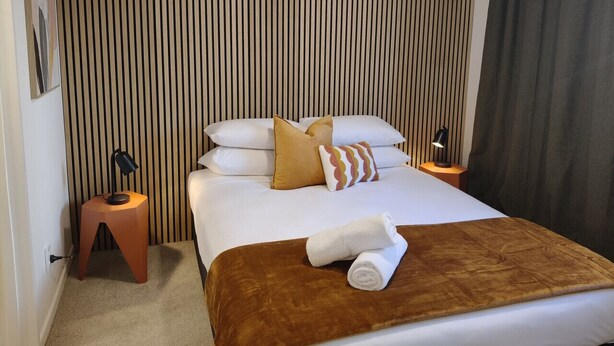 Ein Hotelzimmer mit Bett, Holz-Kopfteil, Nachttisch, Lampe und Handtuch auf dem Bett.