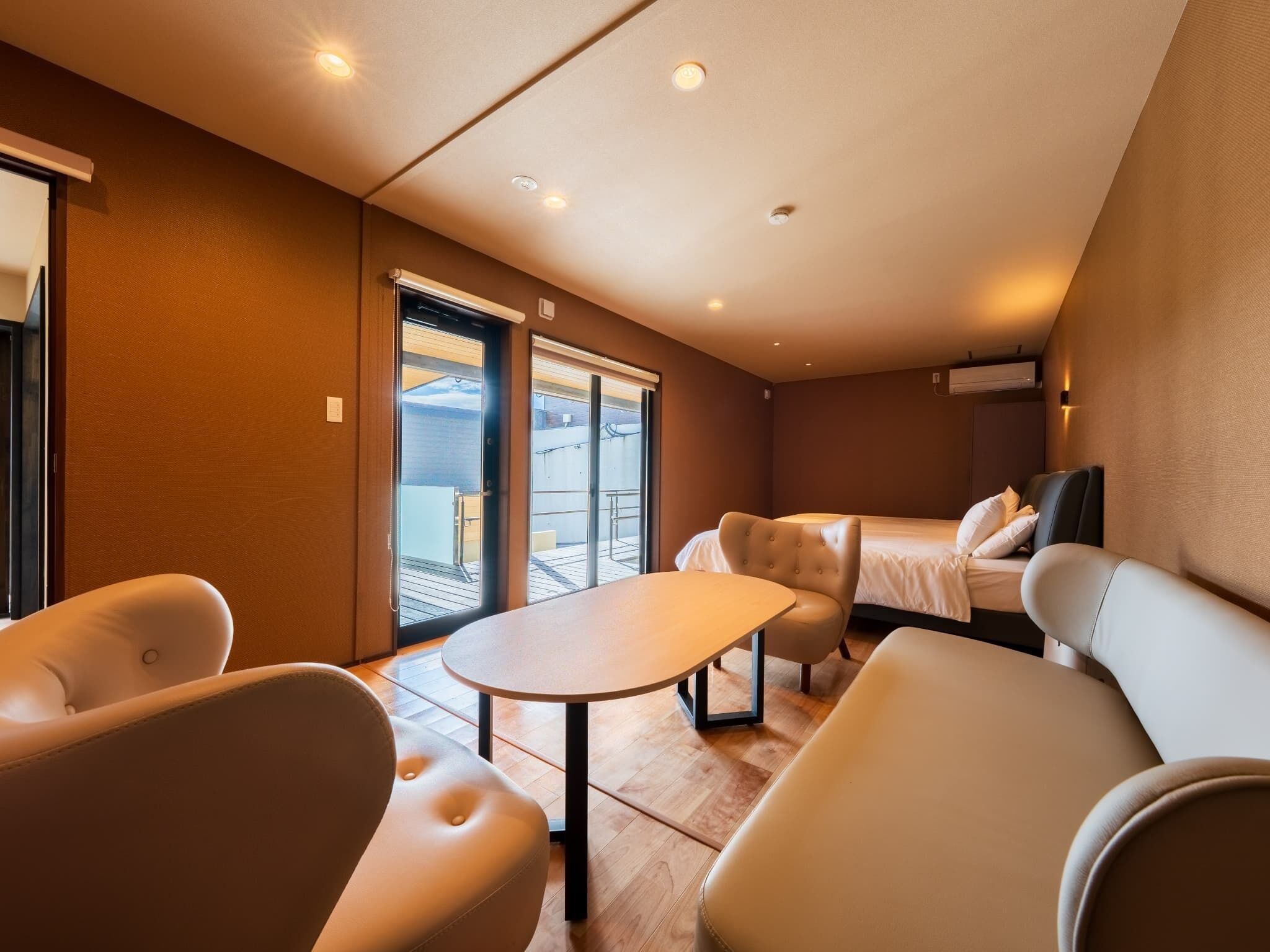 Une chambre d’hôtel moderne équipée d’un lit, de deux fauteuils, d’une petite table et d’une porte-fenêtre coulissante donnant sur un balcon extérieur.