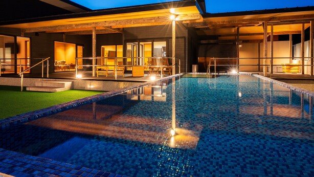Une piscine avec une terrasse carrelée, un espace de détente couvert avec des chaises, et un bâtiment doté de grandes fenêtres.