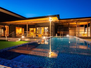 A swimming pool with a tiled deck, a covered seating area with chairs, and a building with large windows. - Umine Annex (Beppu)