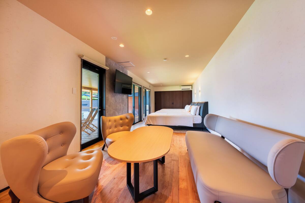 Une chambre d’hôtel moderne, équipée d’un lit, d’un canapé, d’une chaise et d’une table. Une grande fenêtre offre une vue sur l’extérieur.