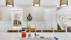 Una cocina con encimera blanca, un lavamanos y una mesa de comedor de madera con sillas. Se observa un ancla decorativa en la pared y una escalera que lleva a otro nivel.