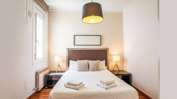 A hotel room with a large bed, two bedside lamps, a framed picture above the bed, and a window on the left. - A3IND1123- Ático near Sagrada Familia (Barcelona)
