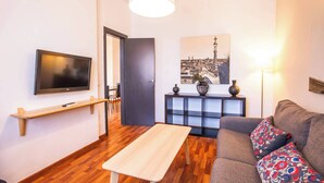 A modern living room with a flat-screen TV, a wooden coffee table, a grey sofa with patterned cushions, a black console table with decorative items, and a cityscape painting on the wall. - A3IND1123- Ático near Sagrada Familia (Barcelona)