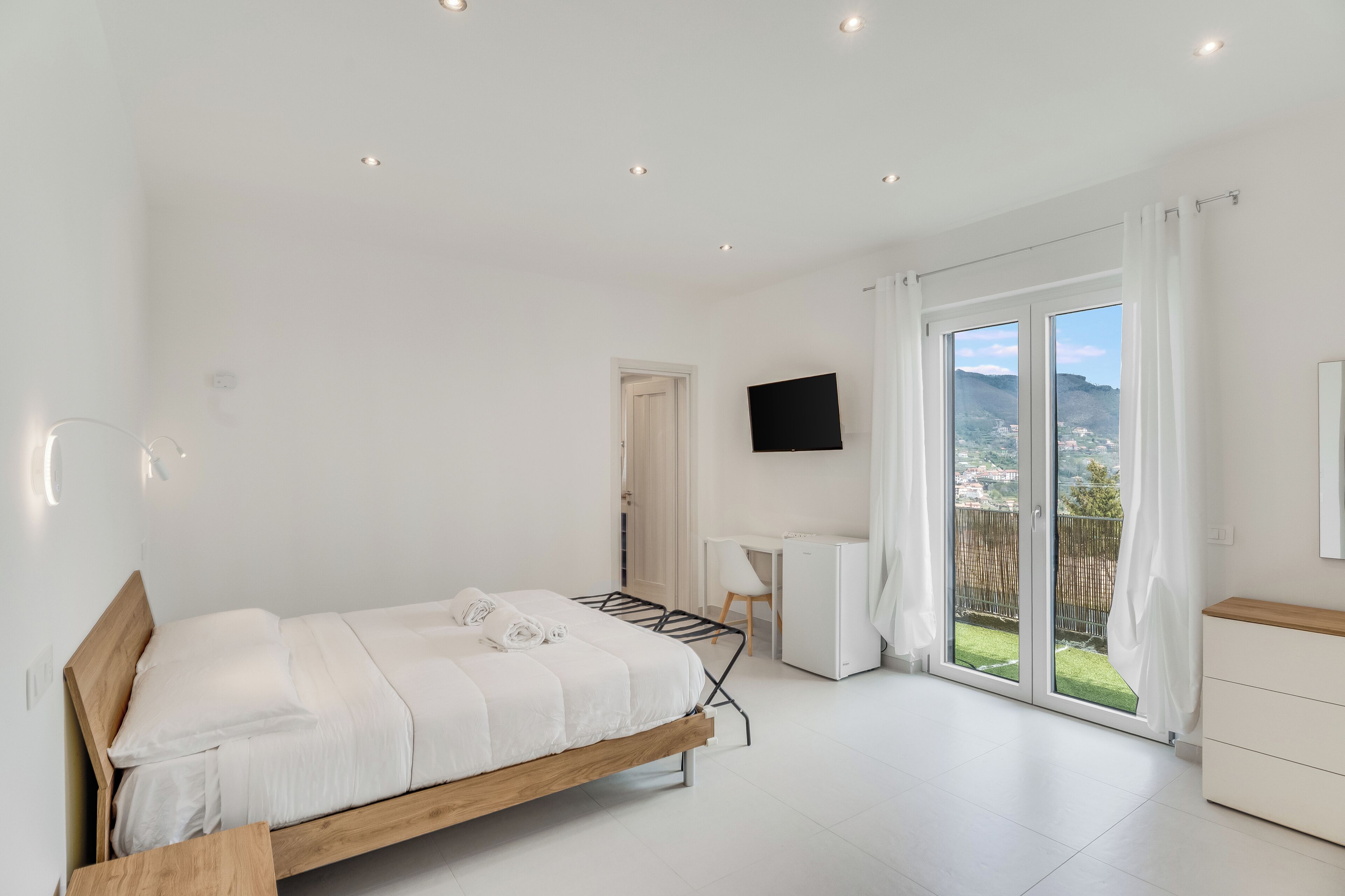Un dormitorio moderno con cama, escritorio, televisor y puerta corrediza de cristal a un balcón.
