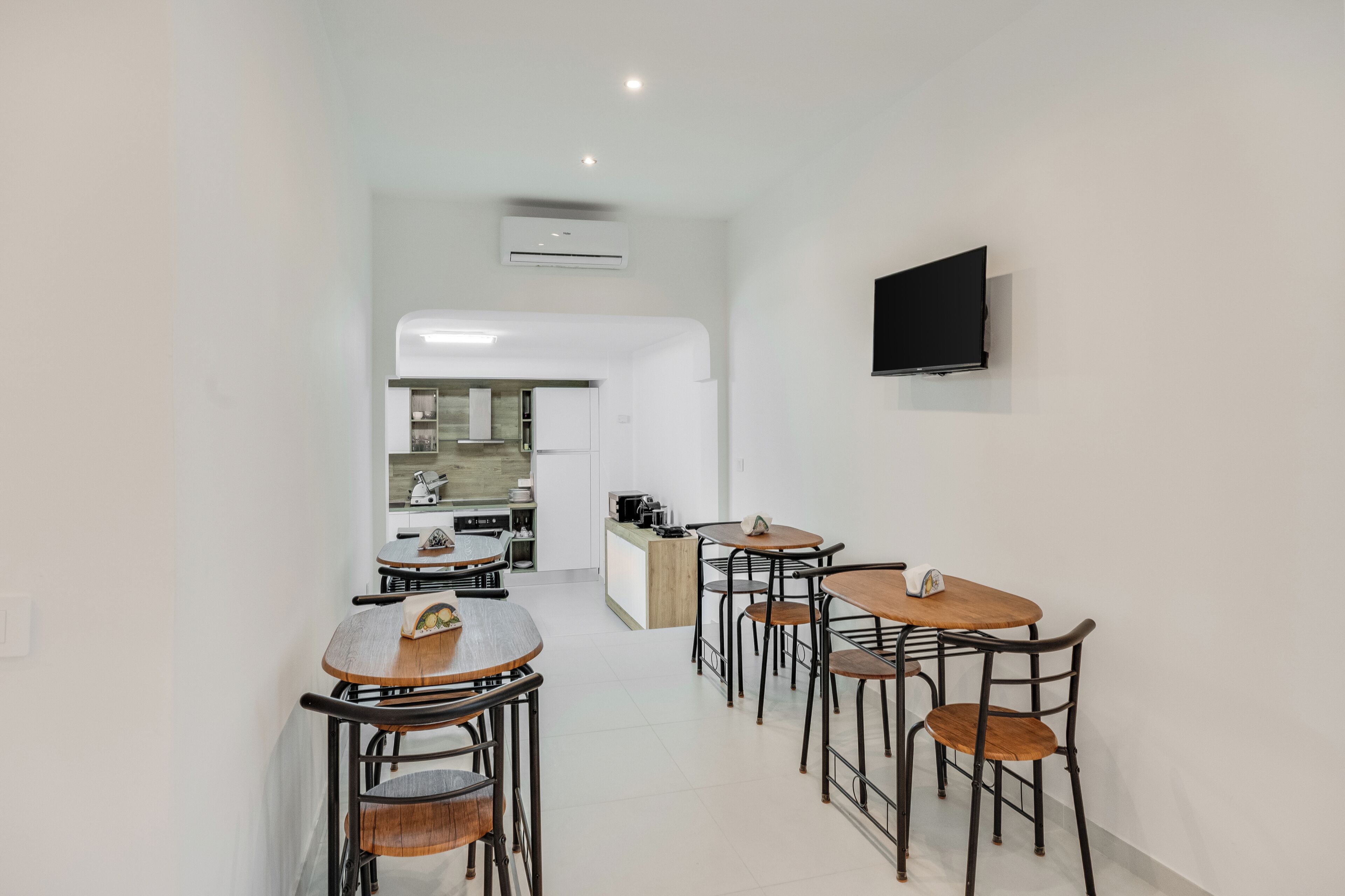 A modern kitchen with white cabinets, a built-in oven, and a microwave. There are two dining tables with chairs, and a flat-screen TV mounted on the wall.
