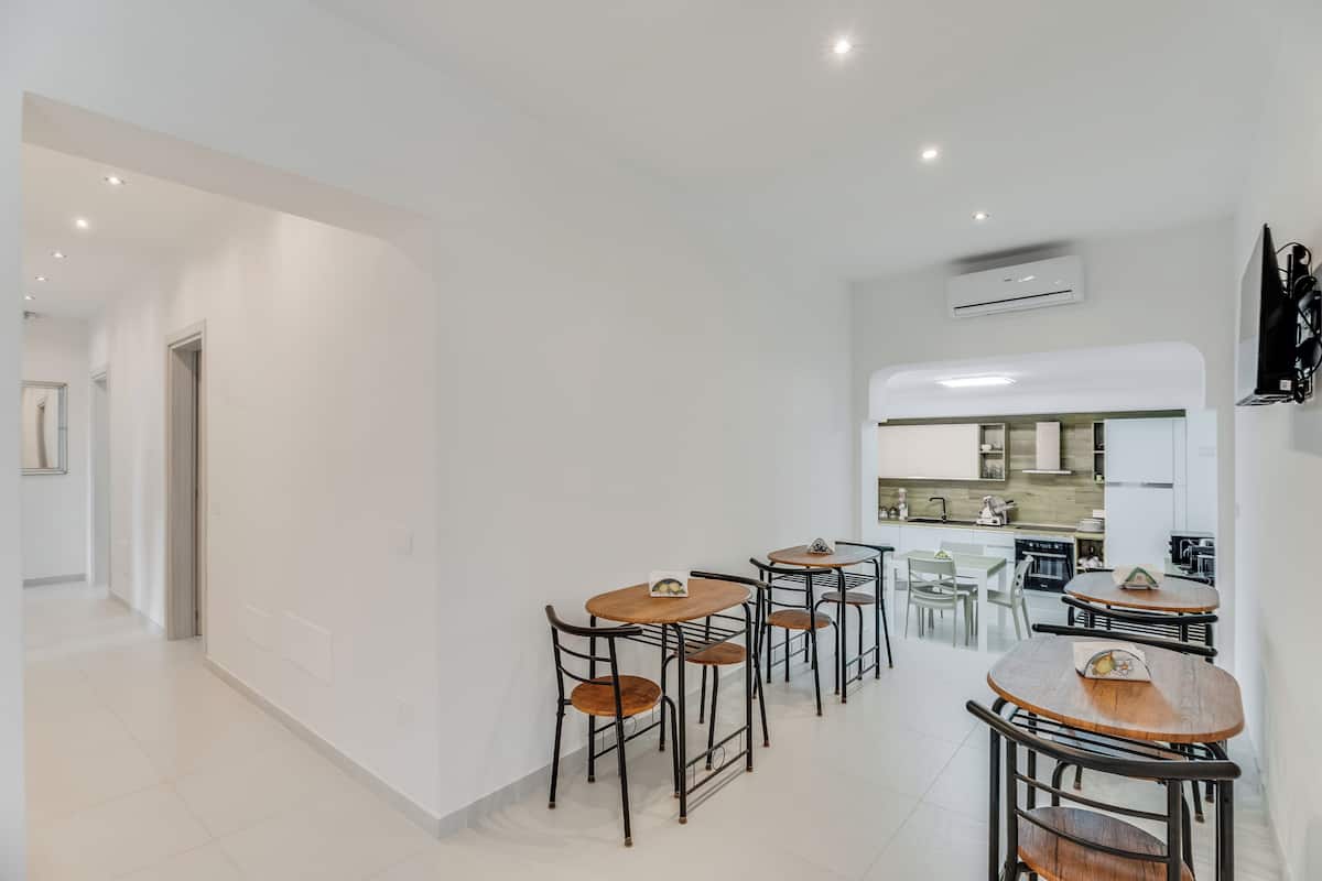 Une cuisine moderne avec des meubles blancs, un coin repas doté de tables et de chaises en bois, et un couloir avec une porte.