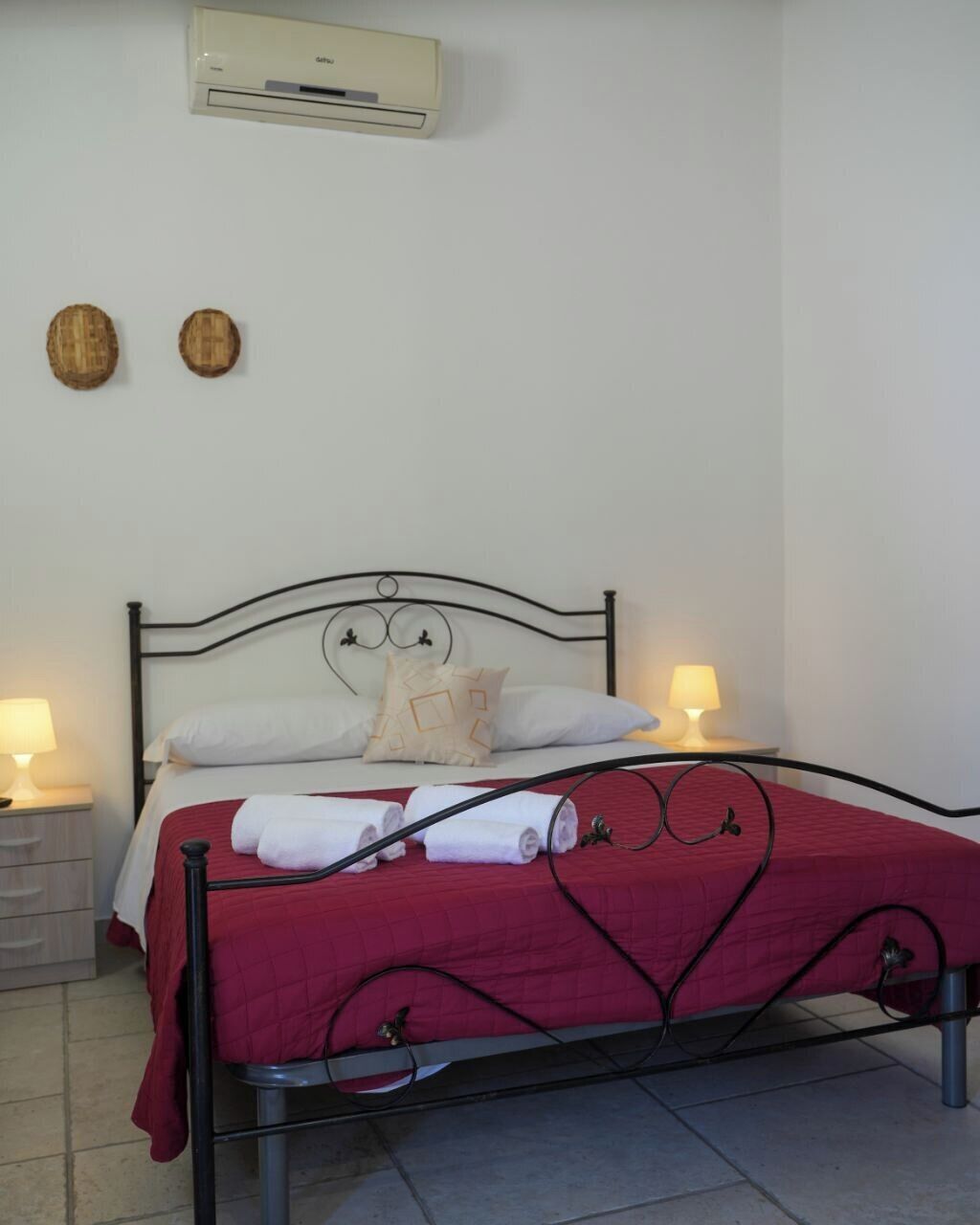 Ein Bett mit roter Steppdecke, zwei Nachttischlampen, eine Wandklimaanlage und zwei Holzdeko-Elemente an der Wand.