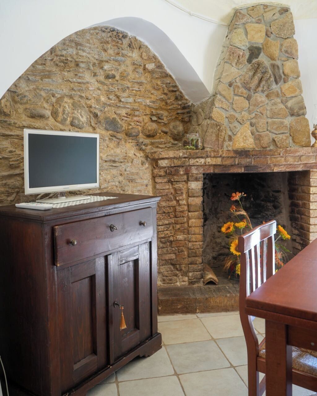 A stone fireplace with a television on a wooden cabinet, a chair, and a vase with flowers.