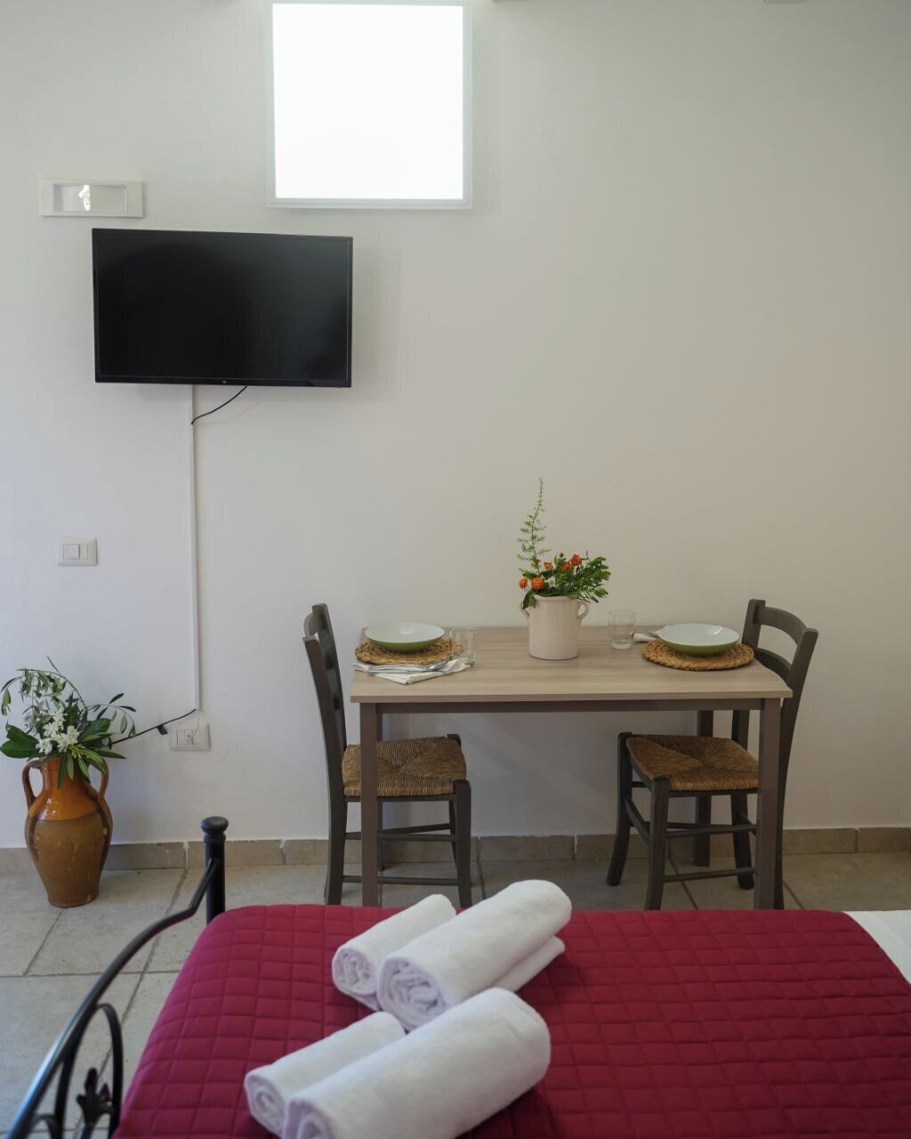 Habitació d'hotel amb un llit, un televisor fixat a la paret, una taula de menjador amb cadires i una petita planta en test.