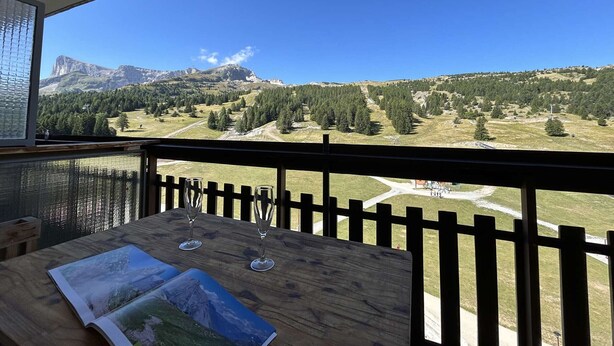 Una mesa de madera con dos copas de vino y un libro, con vistas a un paisaje montañoso y cielo despejado.