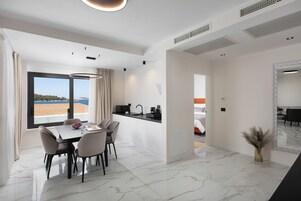 A modern hotel room with a dining area, a kitchen, and a large window offering a view of the sea.