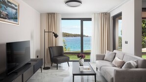 Una sala de estar moderna con un televisor de pantalla plana grande, un sofá, una silla y una mesa de centro. Hay una ventana con vistas al mar y al paisaje.