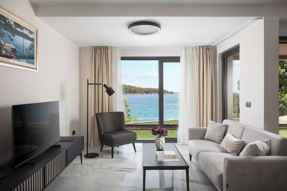 A modern living room with a large flat-screen TV, a sofa, a chair, and a coffee table. There is a window with a view of the sea and a landscape.