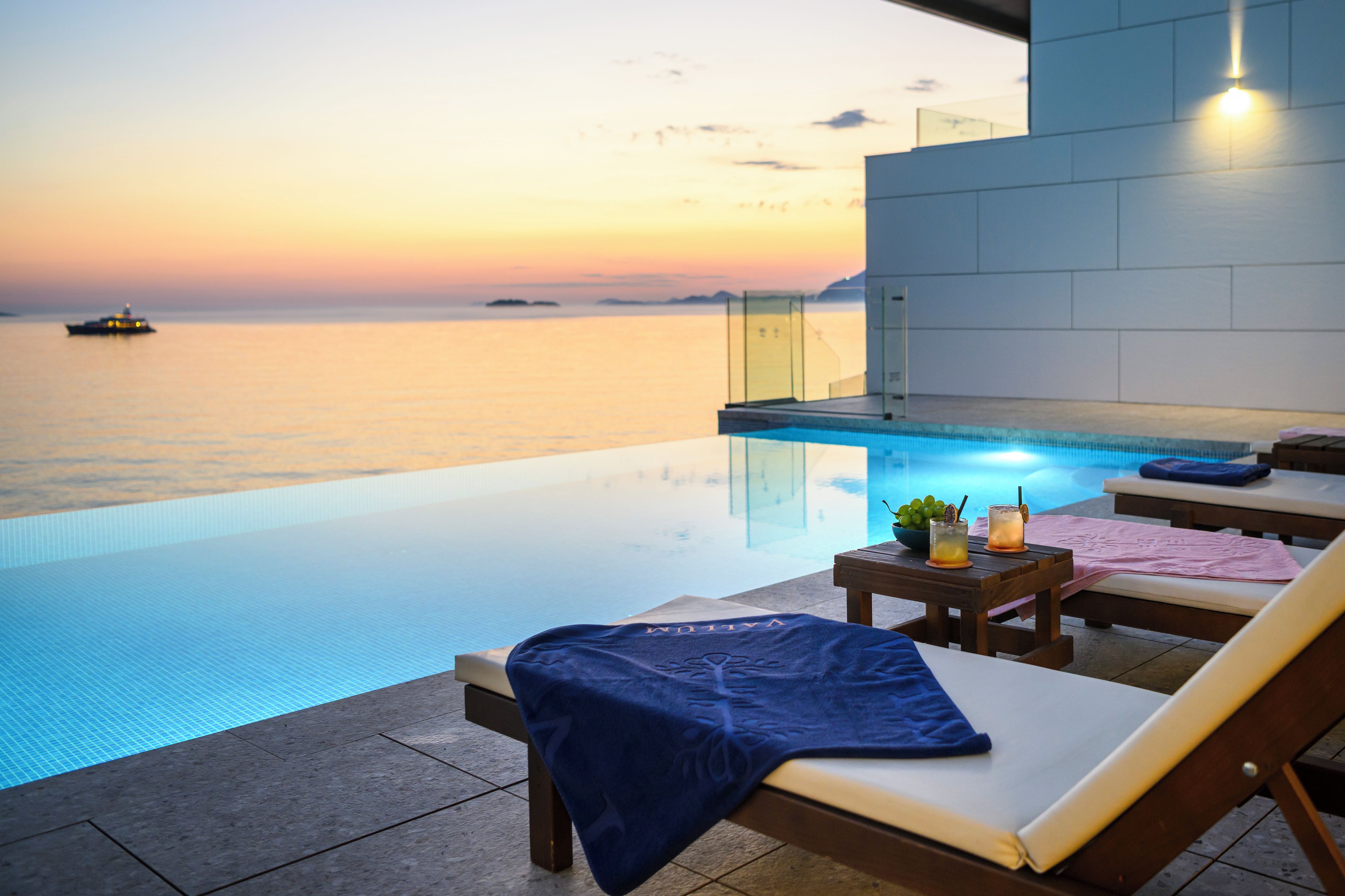 Piscina infinita con vista despejada al mar y un atardecer. Hay sillones de descanso y una mesa pequeña con bebidas.