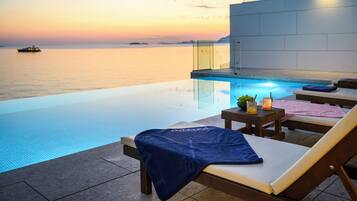 Piscina infinita con vista despejada al mar y un atardecer. Hay sillones de descanso y una mesa pequeña con bebidas.