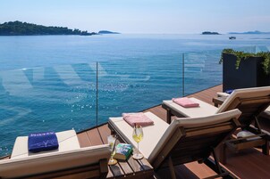 Una terraza con sillones de descanso, un vaso de bebida y un libro, con vista a una masa de agua con barcos e islas distantes.