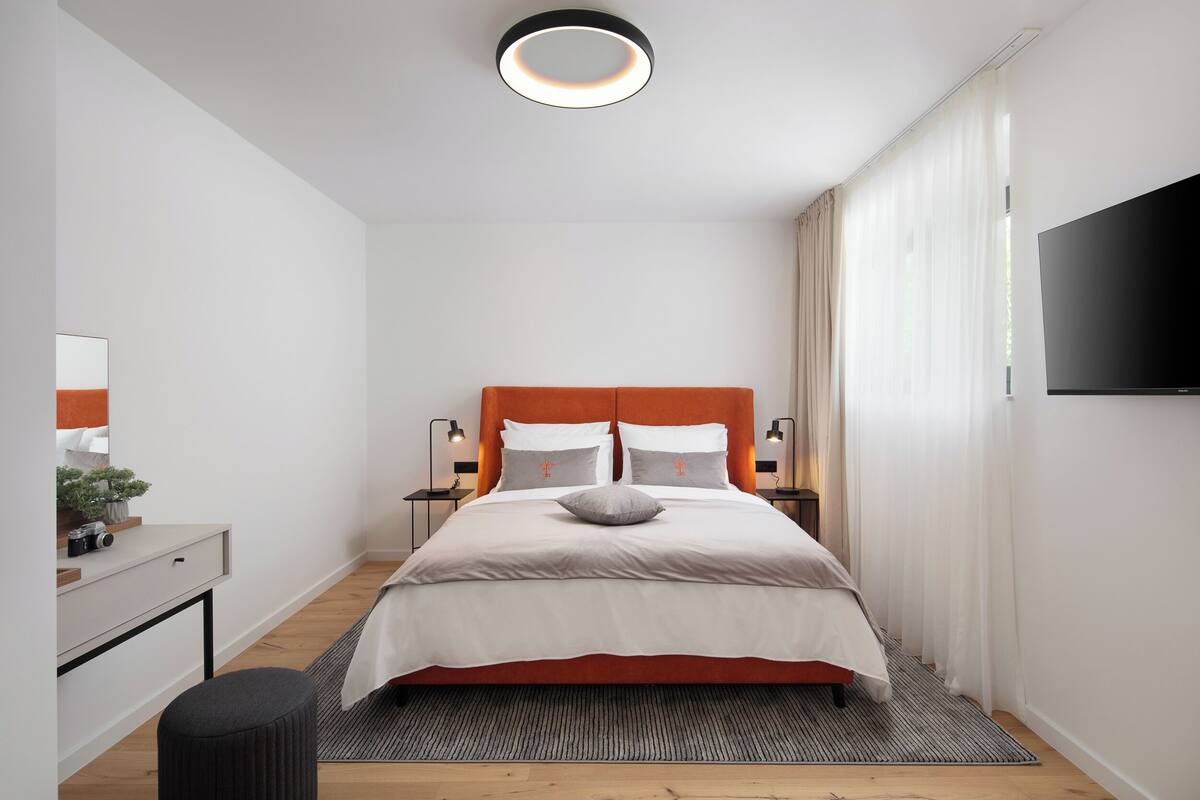 A modern bedroom with a large bed, bedside lamps, a flat-screen TV, a desk, and a mirror.