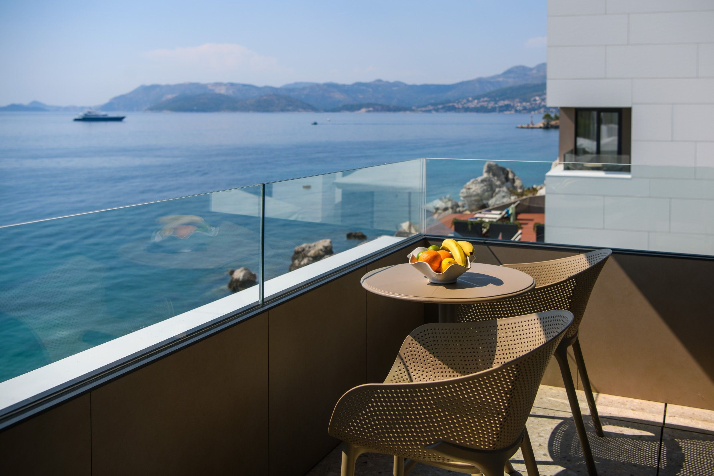 Un balcon avec vue sur la mer et les montagnes, une table avec un panier de fruits et des chaises en osier.
